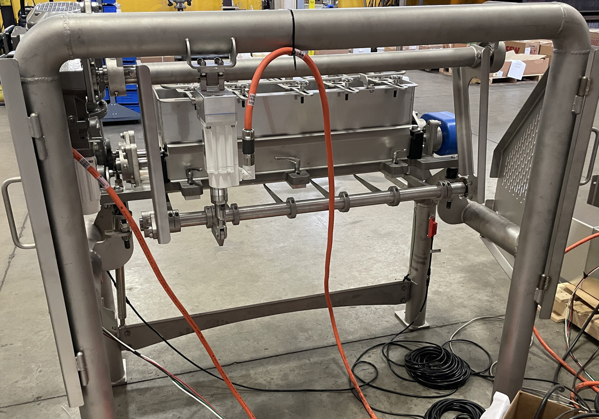 Depositor machine for food processing with stainless steel construction, featuring hoses and connections for material handling, set in a warehouse environment.